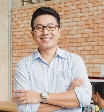 Smiling man in a casual shirt standing with folded arms in an office setting with a brick wall background.