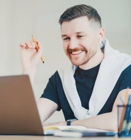 Man smiling while working on a laptop, holding a pen, wearing earphones and a sweater draped over shoulders.