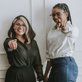 Two people smiling and pointing at the camera in a friendly, confident manner, standing against a white wall background.